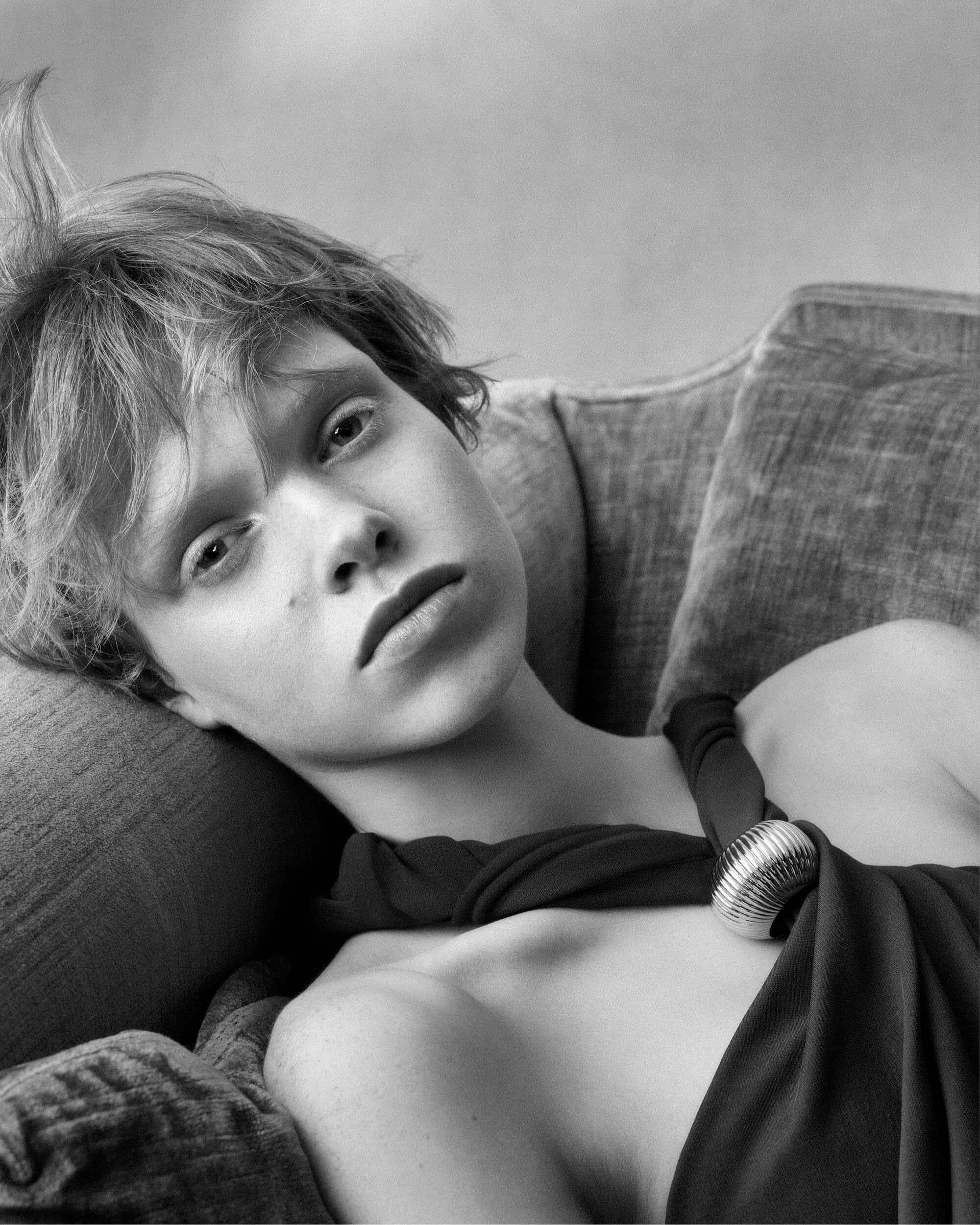 Monochrome portrait of a model resting on an upholstered sofa wearing a dark garment with a metallic accessory.
