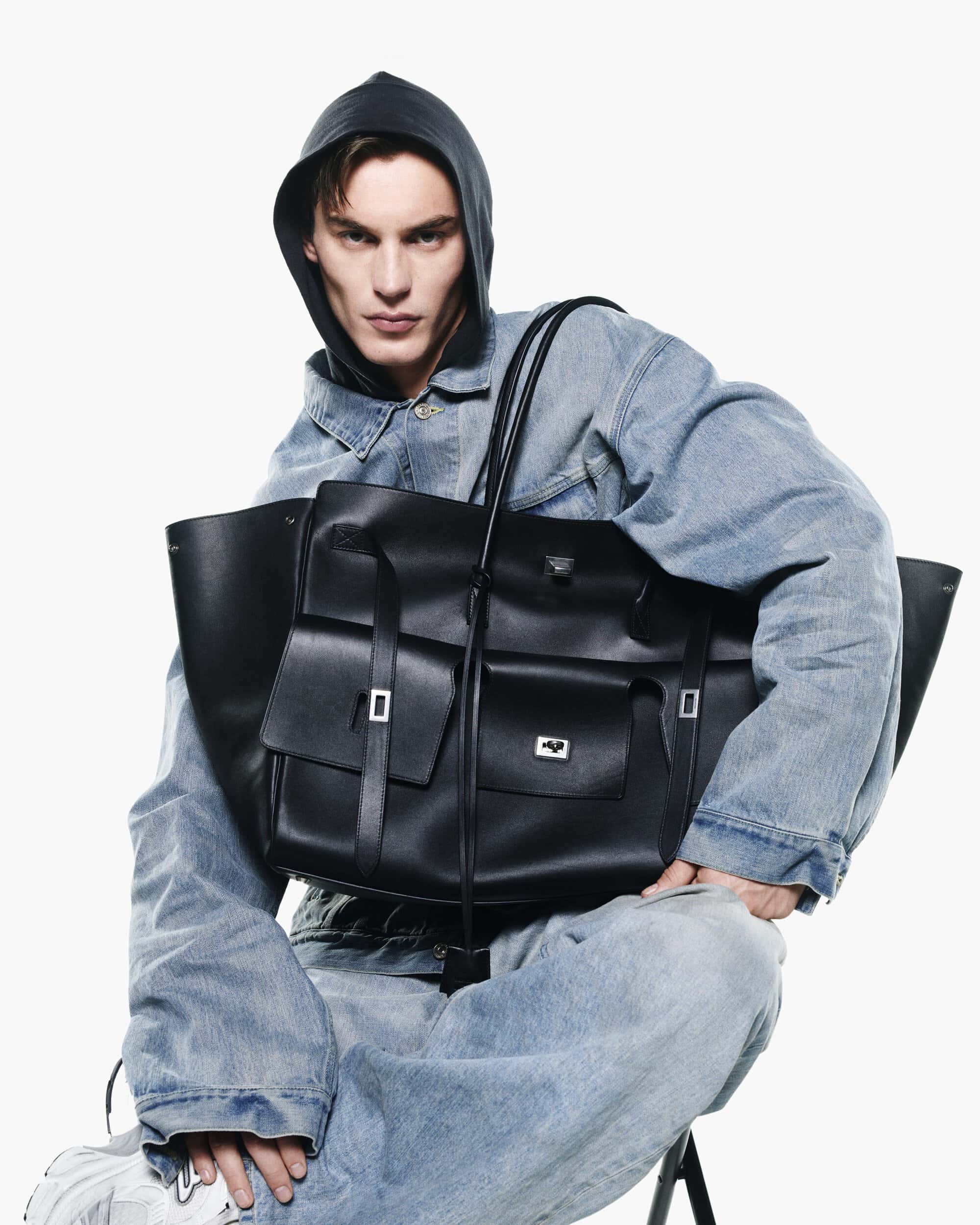 Man wearing a denim jacket and hooded sweatshirt holding a large black leather bag against a white background.
