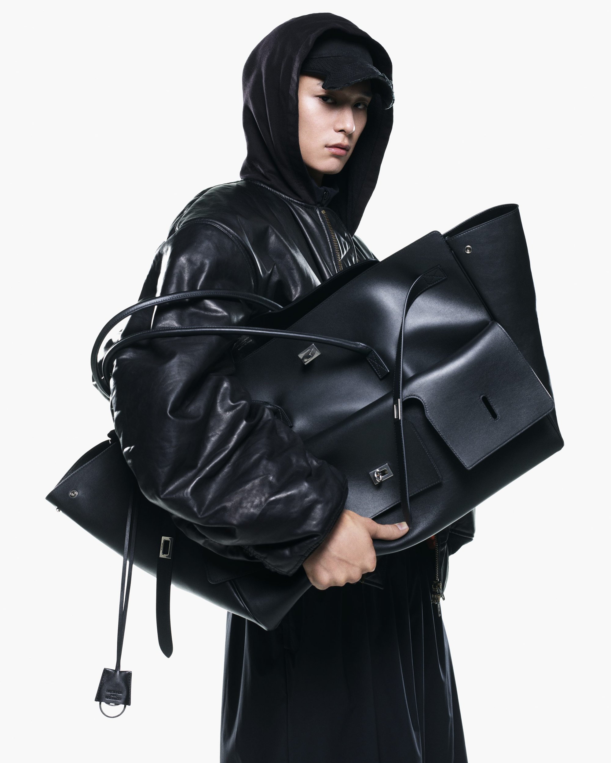 Man wearing a black leather jacket and hoodie holding multiple large black leather tote bags against a white background
