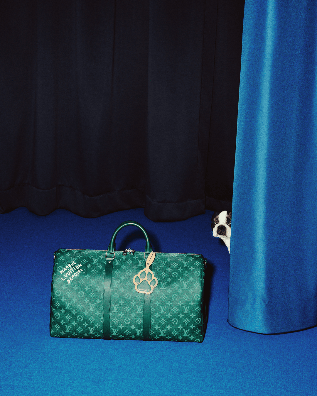 Louis Vuitton green monogram duffel bag sitting on blue floor with a small dog peeking from behind a curtain