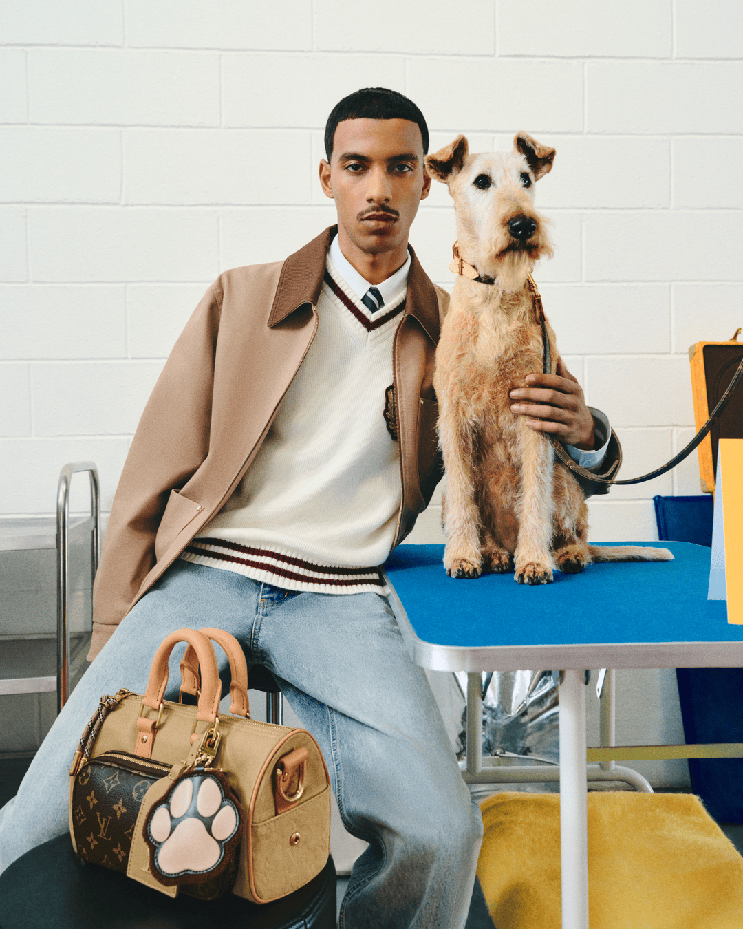 Model seated beside a wirehaired terrier on a table, holding a monogram handbag featuring a paw-shaped charm.