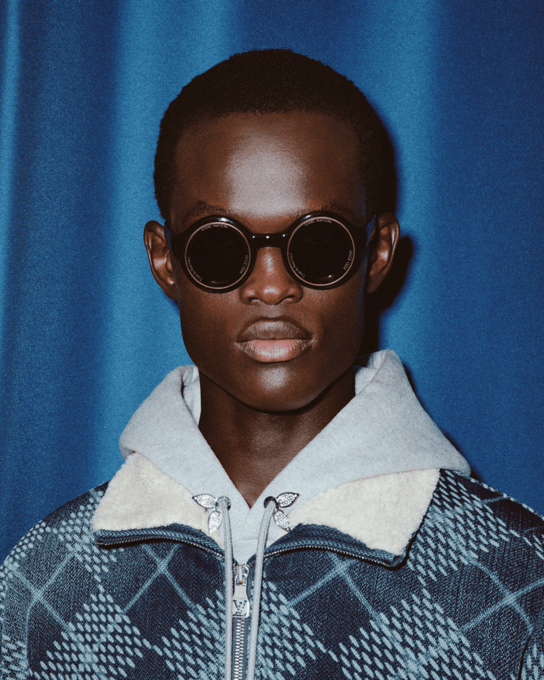 Portrait of a man wearing round sunglasses and a plaid jacket against a blue textured curtain background