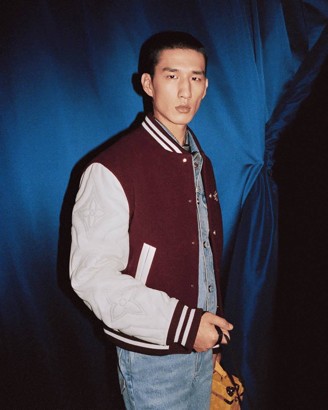 Model wearing a burgundy varsity jacket with white sleeves and a denim shirt against a deep blue curtain background.