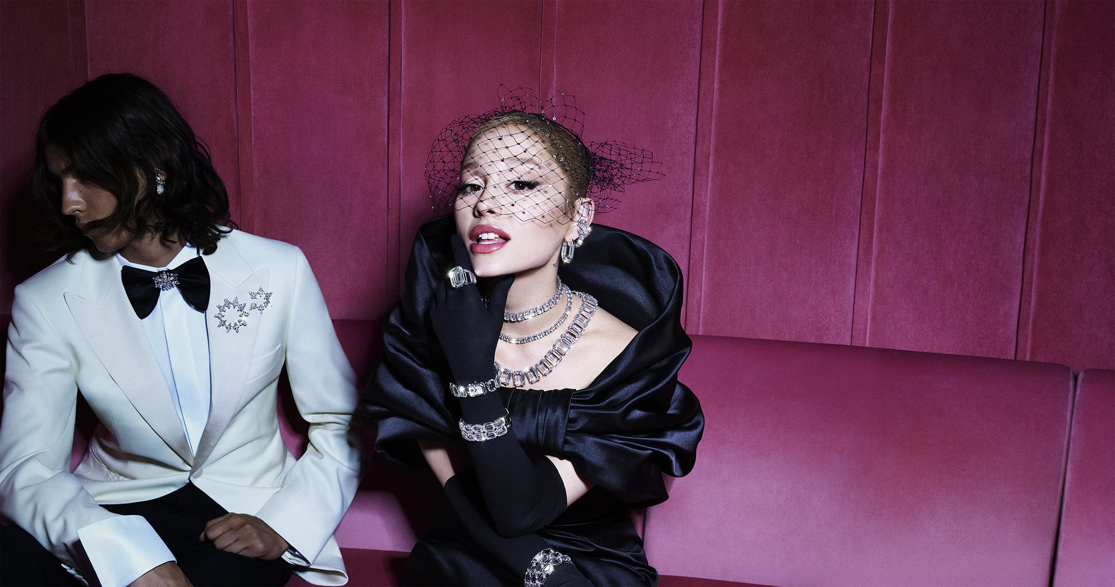Ariana Grande in a black dress with crystal jewelry next to a man in a white tuxedo jacket on a pink sofa