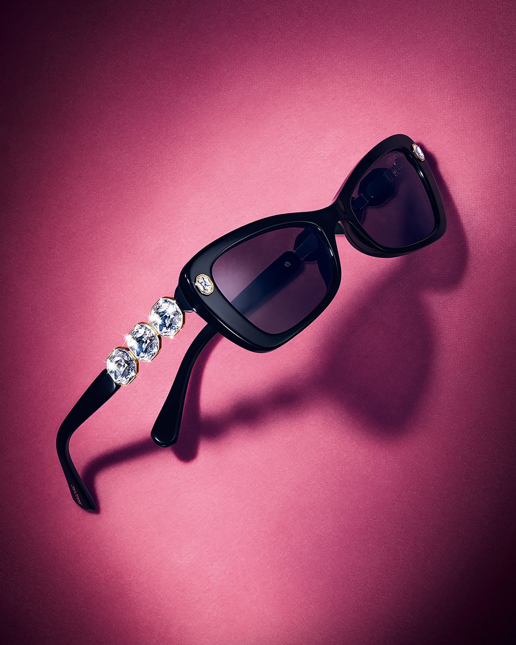 Black sunglasses with crystal-embellished temples resting against a rich pink textured background