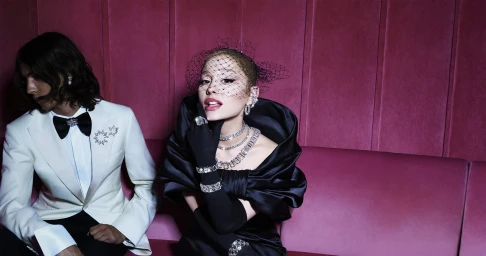 Ariana Grande in a black dress with crystal jewelry next to a man in a white tuxedo jacket on a pink sofa