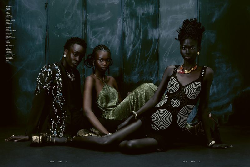Three dark-skinned fashion models pose indoors against a dark wall wearing modern dresses and bold gold jewellery in a studio shot.