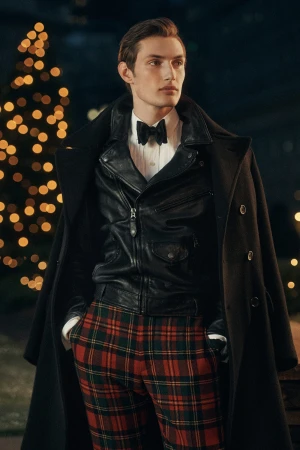 Man wearing a black leather jacket and tartan trousers under a heavy dark coat with a festive light background