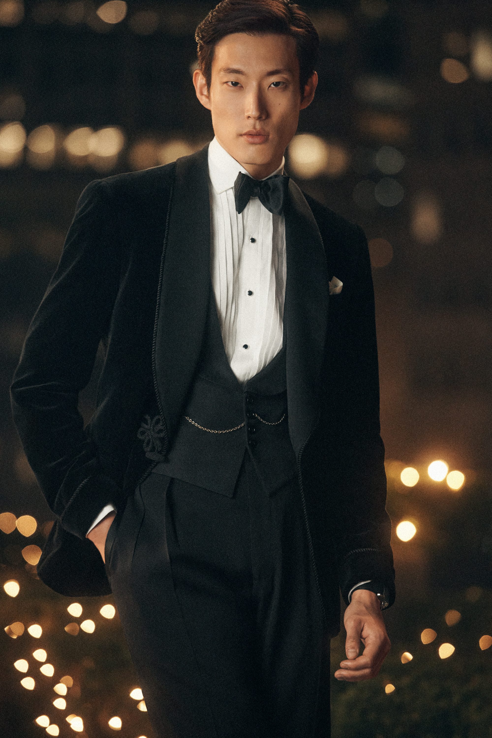 Man wearing a black velvet tuxedo jacket, white pleated shirt, and bow tie against a softly blurred city night background.