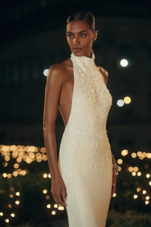 Woman in a white embellished backless halter gown posing outdoors at night against out-of-focus golden lights.