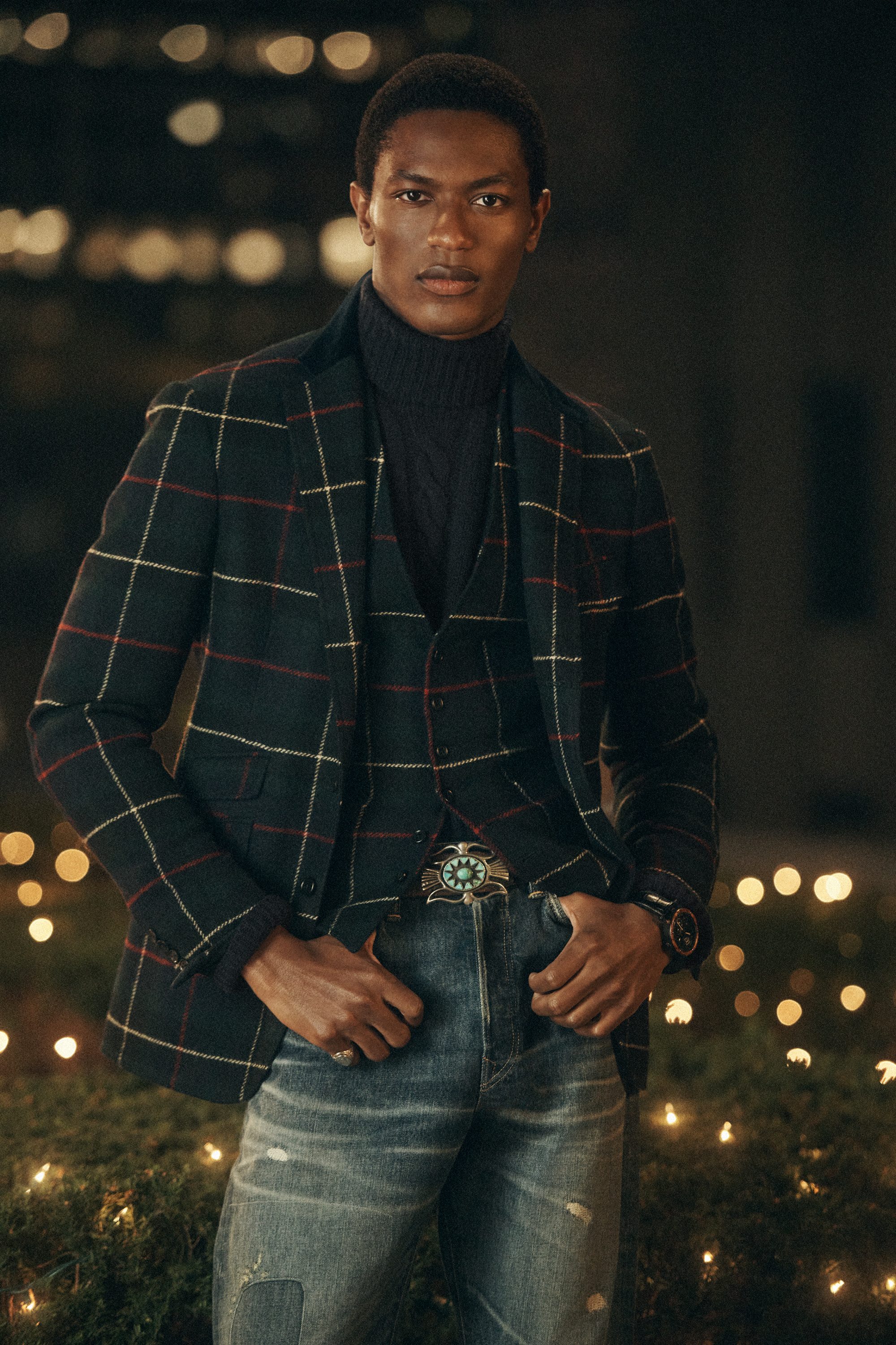 Black man wearing a plaid blazer and turtleneck sweater outdoors at night with bokeh city lights