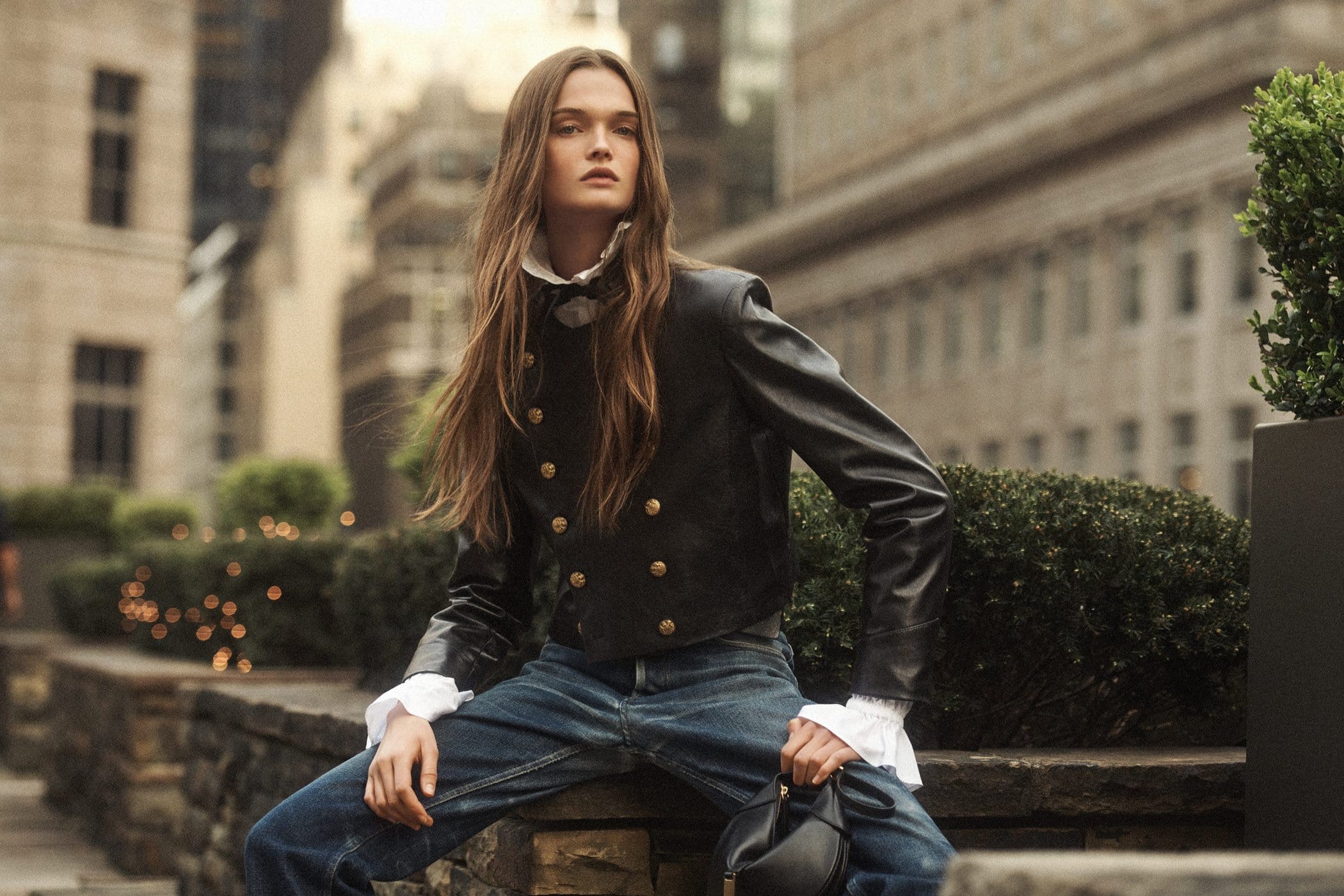 Model in a black leather jacket and blue jeans sitting outdoors against an urban street background with greenery.