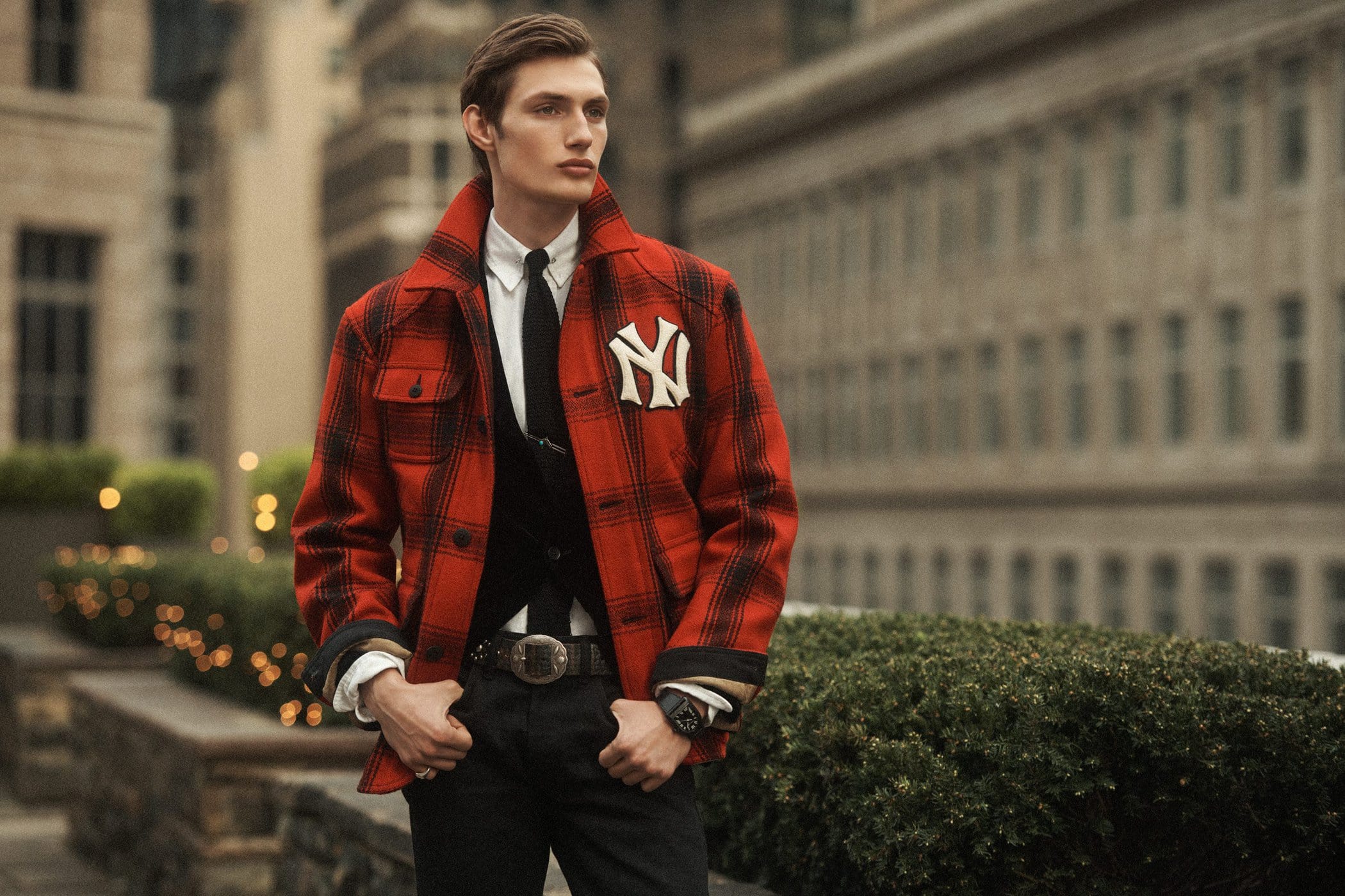 A man wearing a red plaid jacket with a white NY logo standing outdoors against a city building backdrop