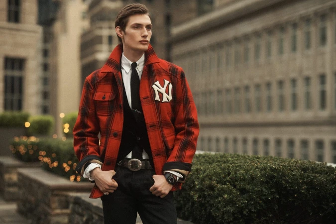 A man wearing a red plaid jacket with a white NY logo standing outdoors against a city building backdrop