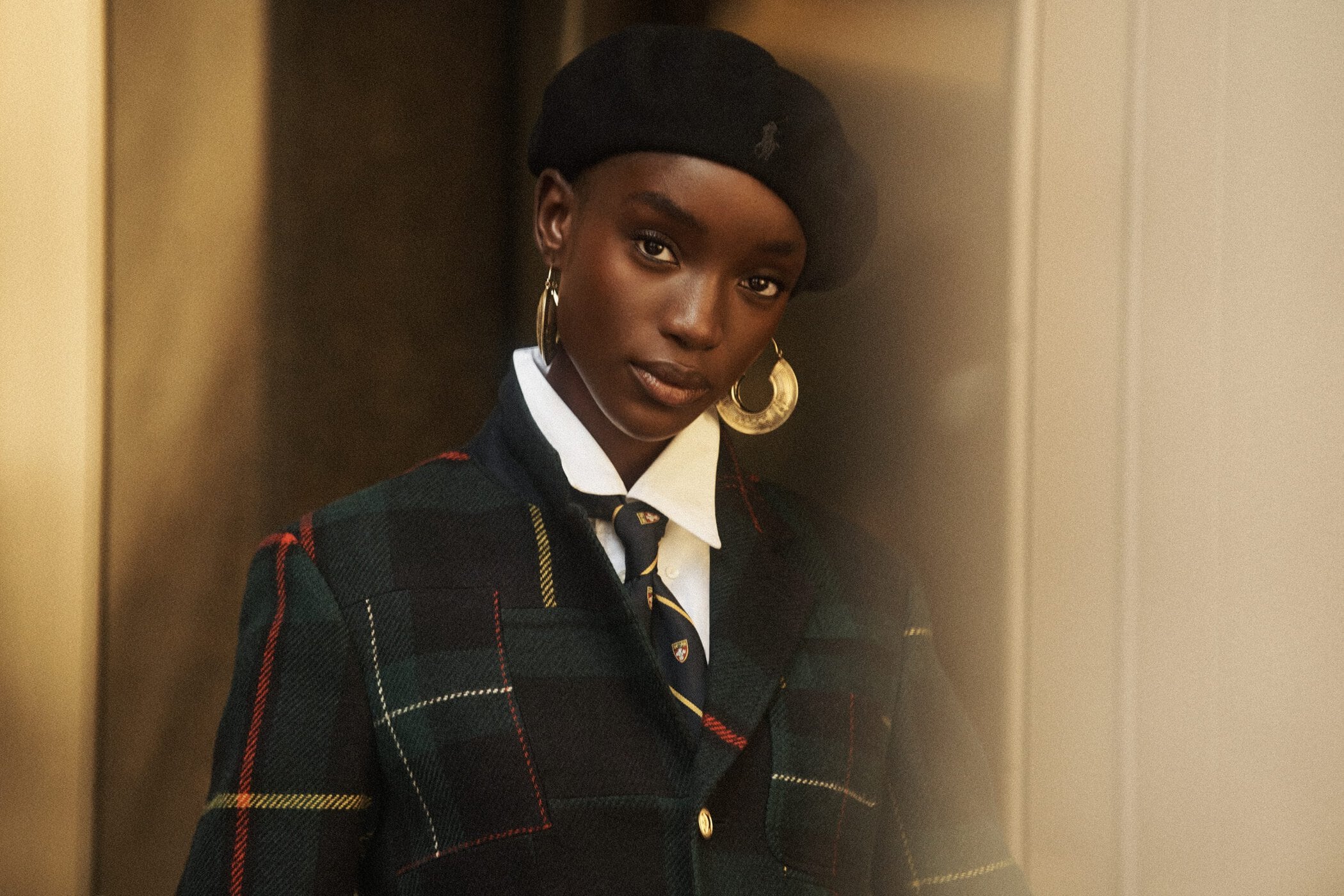 Model wearing a dark tartan blazer, white collared shirt, tie, beret, and gold hoop earrings against a warm interior backdrop.