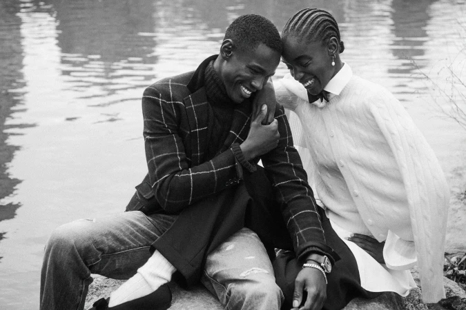 Black and white portrait of a smiling couple sitting outdoors near water, wearing patterned blazers and knitwear.