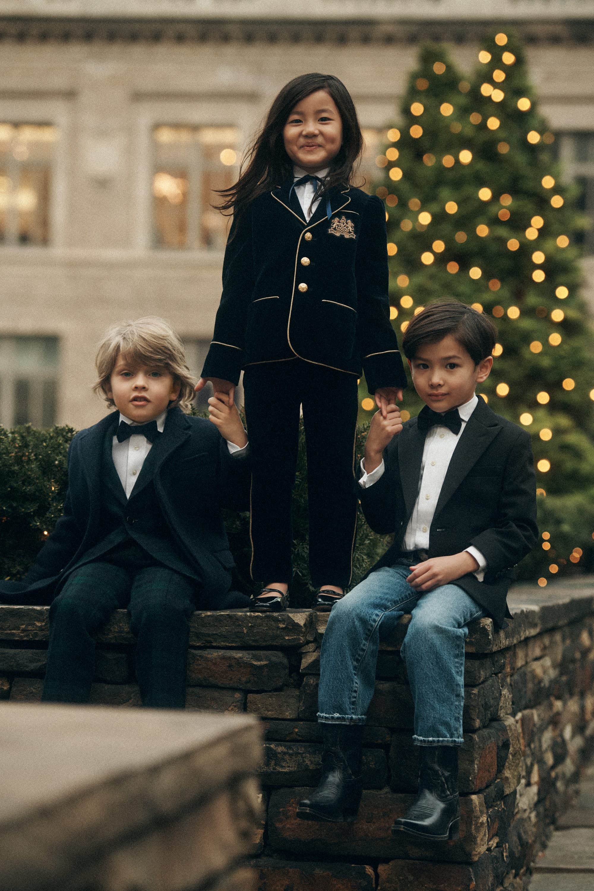 Three children posing outdoors near a holiday tree, wearing formal blazers, bows, and black trousers against a stone wall.