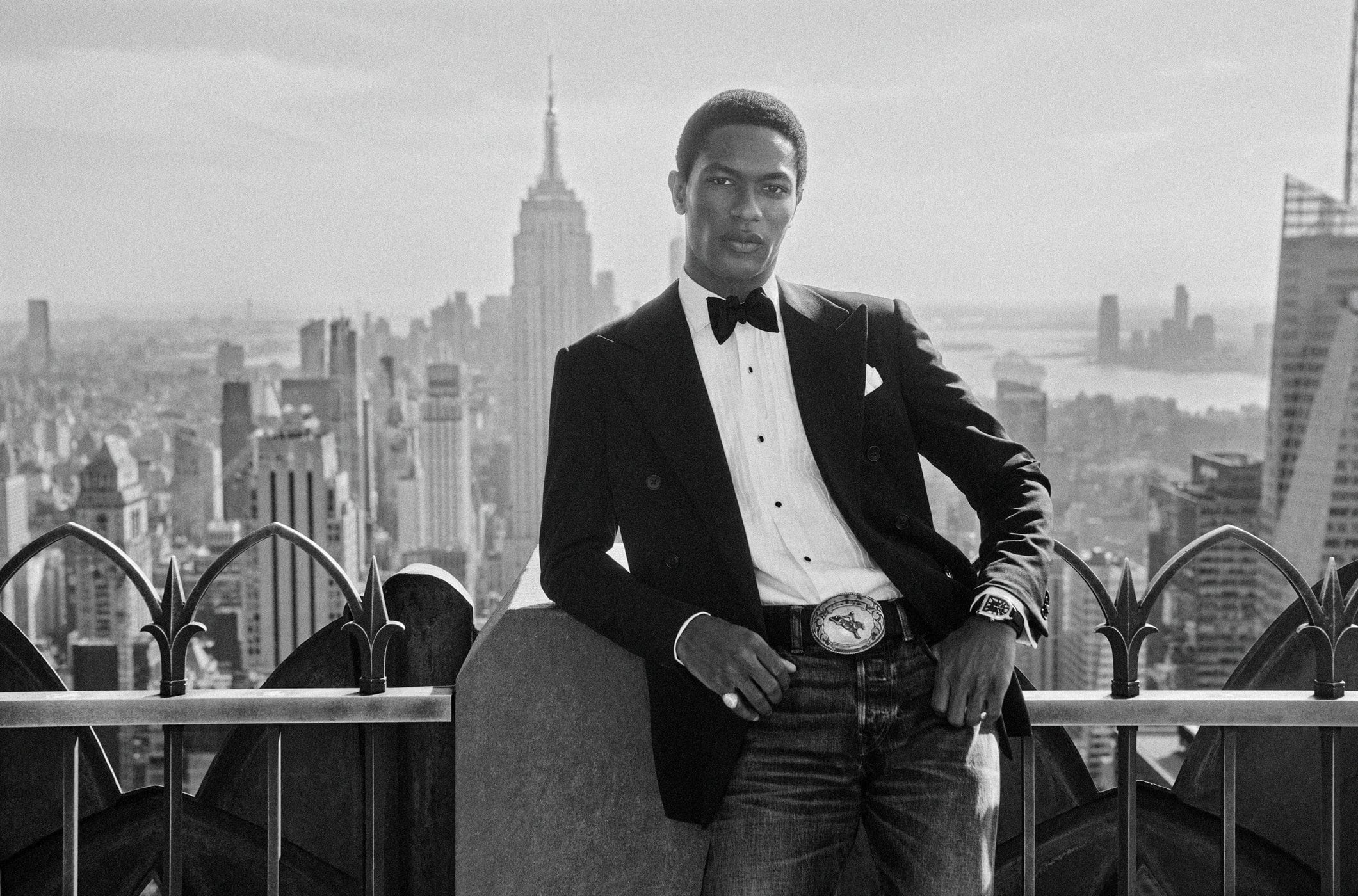 Man wearing a black tuxedo jacket, bow tie, and denim jeans posing on a New York City rooftop overlooking the skyline
