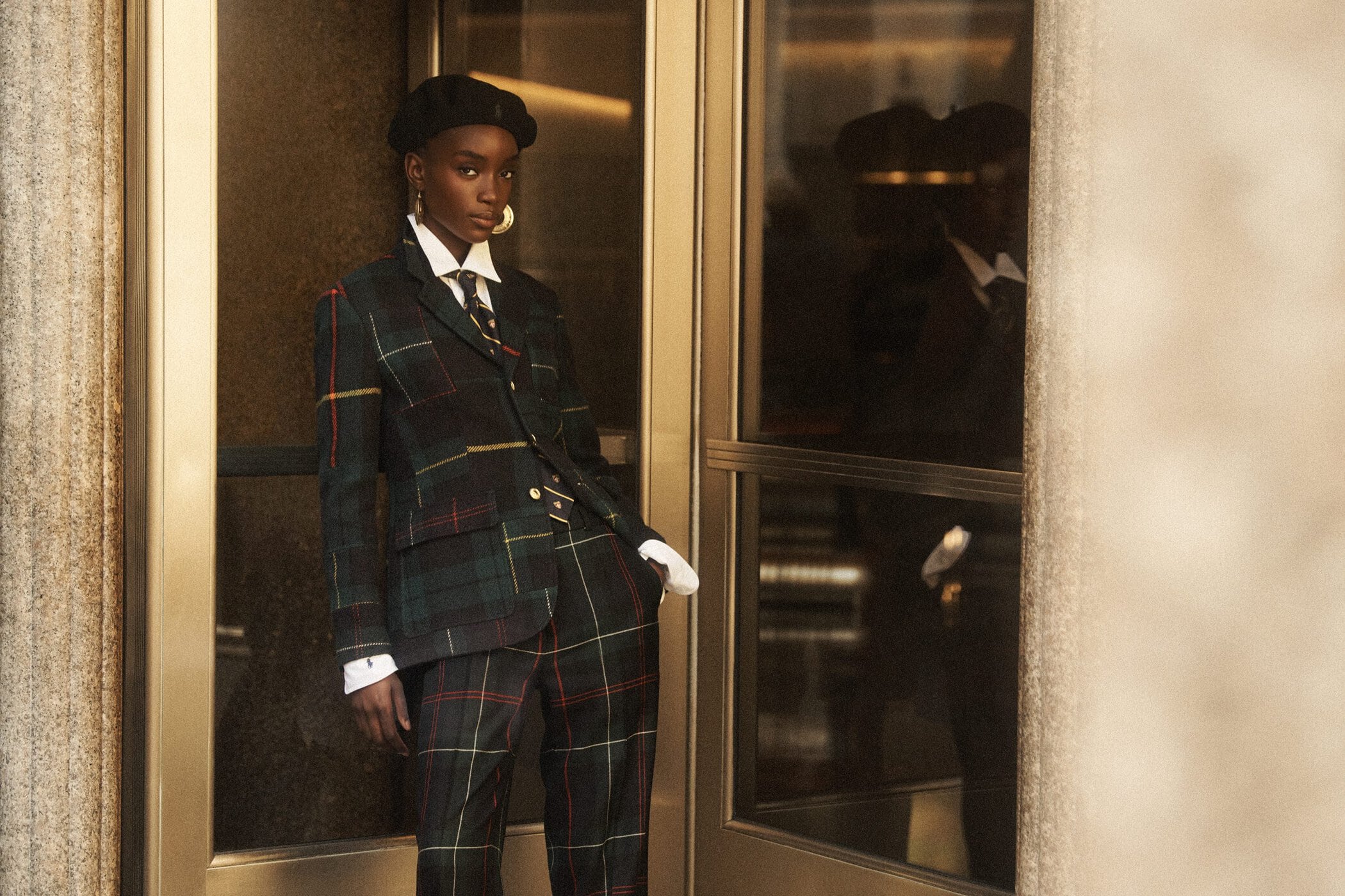 Model wearing a plaid suit, tie, white shirt, and beret standing by a glass entrance for Ralph Lauren