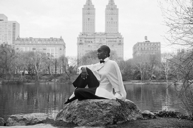 Model seated on a large rock by a lake with New York City architecture in the background, captured in black and white.