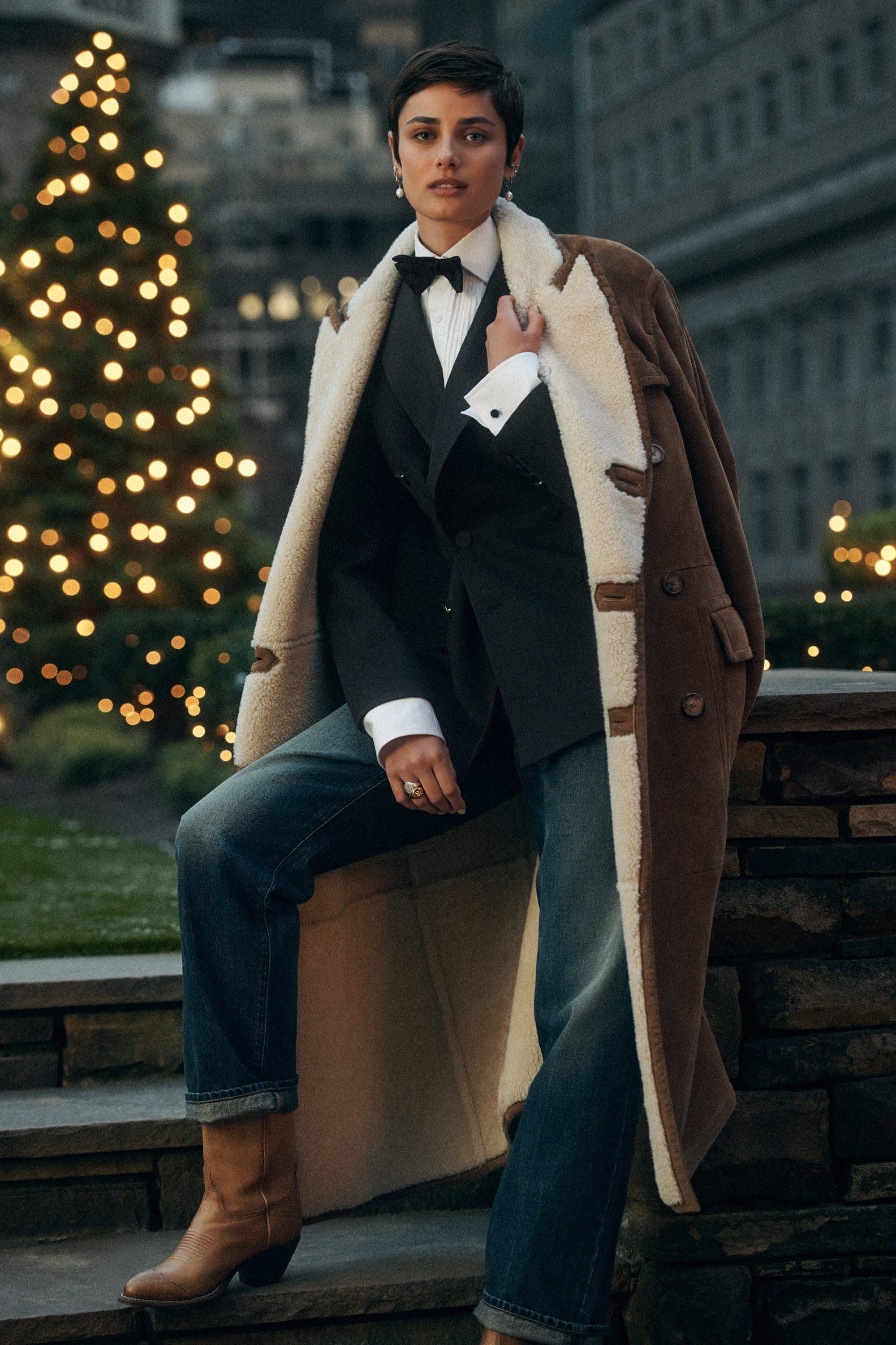 Taylor Hill in a tailored suit and shearling coat outdoors at dusk with twinkling holiday lights.