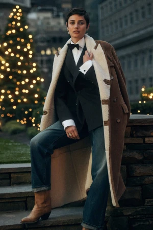 Taylor Hill in a tailored suit and shearling coat outdoors at dusk with twinkling holiday lights.