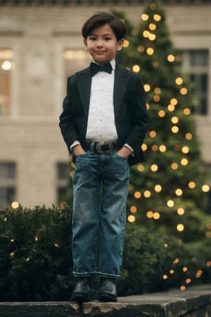 Young boy in a black blazer, bow tie, and denim jeans posing outdoors near a festive illuminated tree