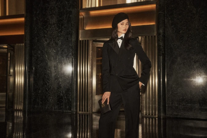 Model wearing black tuxedo and beret holding a clutch in a hotel lobby interior