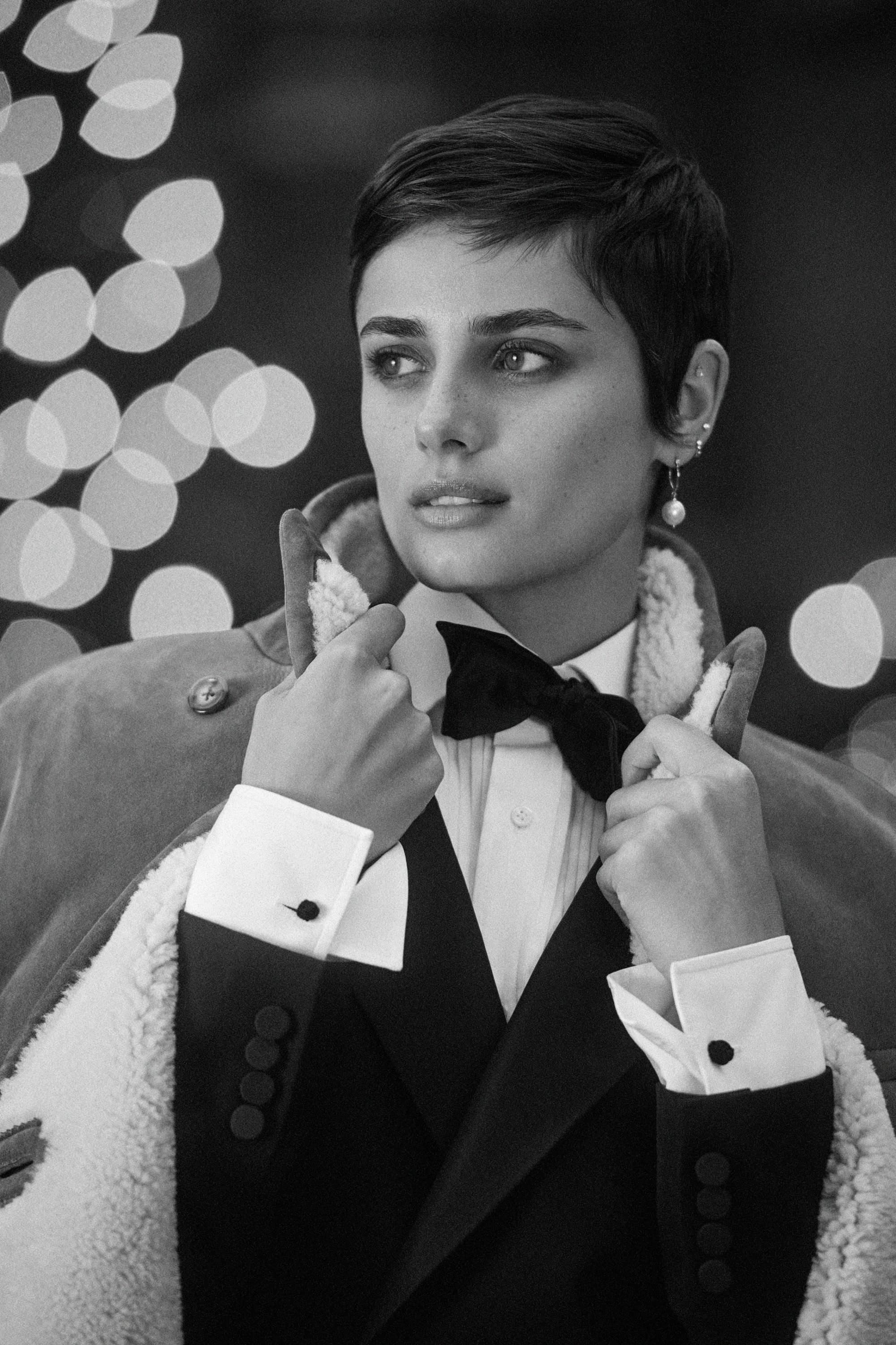 Close-up black and white portrait of a short-haired woman wearing a tuxedo with a bow tie and a shearling-lined coat.