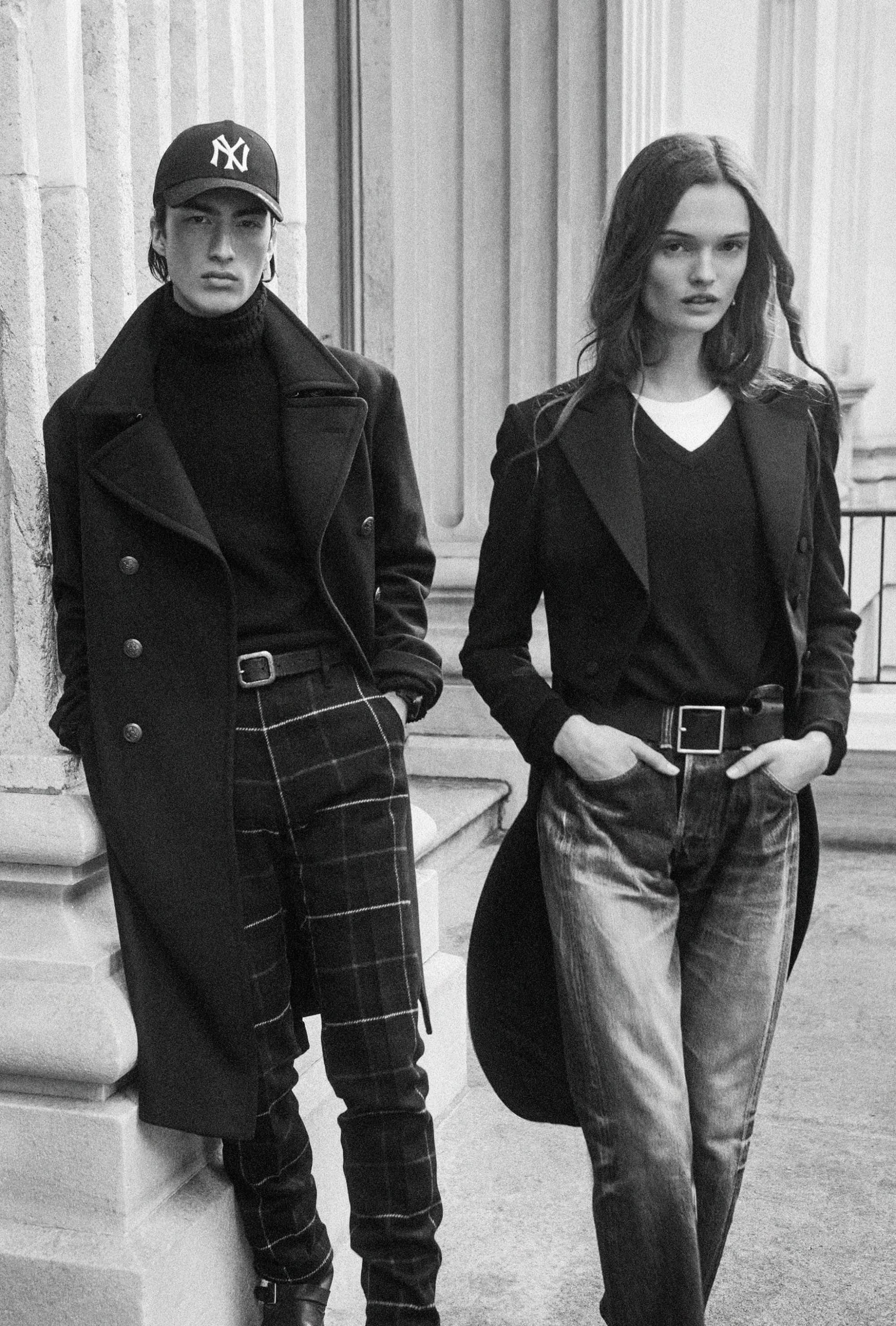 Two fashion models posing against classical architecture in tailored black coats and trousers in a monochrome image