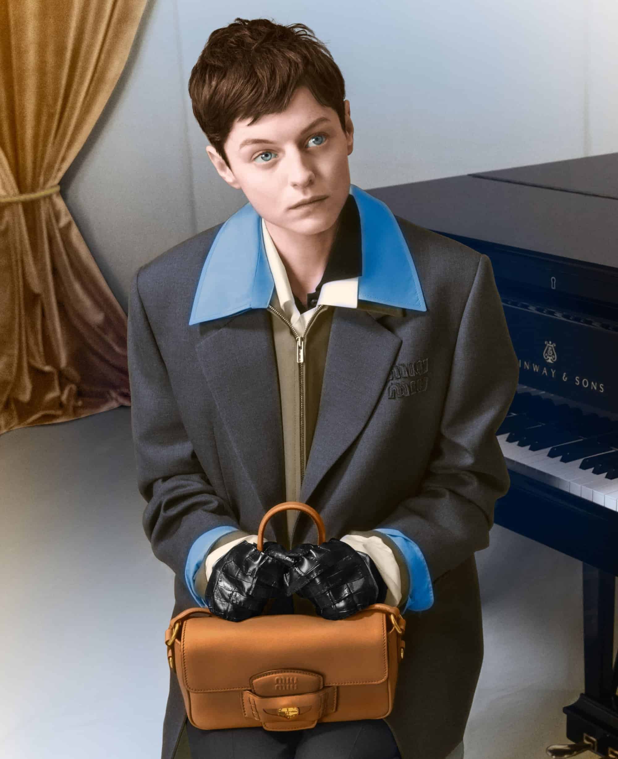 Emma Corrin in grey jacket holding a brown leather handbag with black gloves in a room with a piano