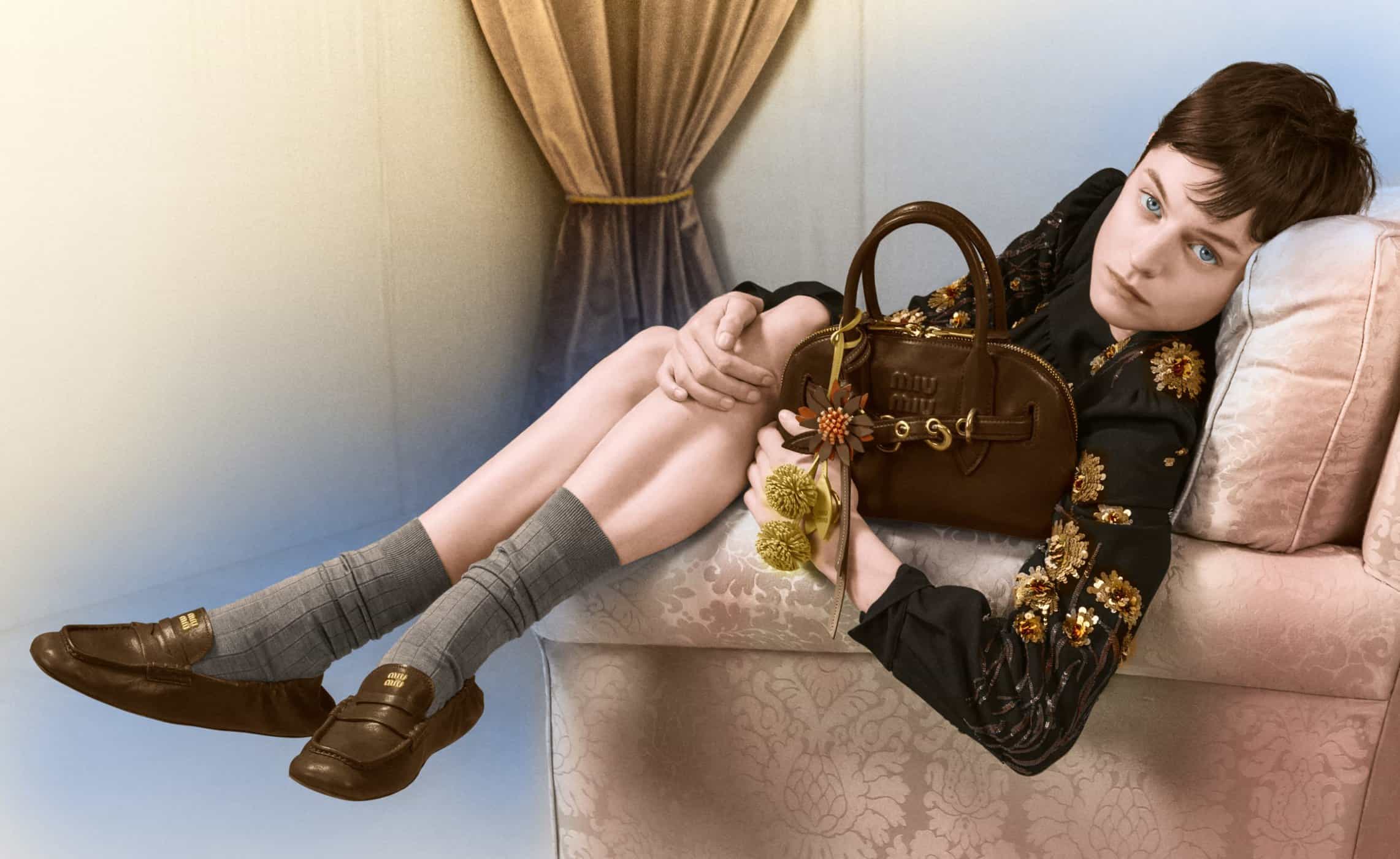 Emma Corrin reclining on a sofa holding a brown Miu Miu handbag for the Holiday 2024 campaign