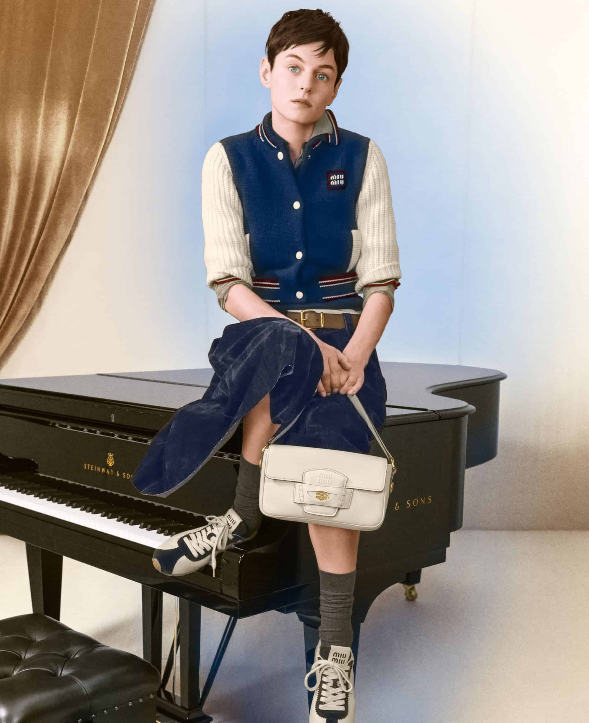 Emma Corrin seated on a black grand piano wearing a blue varsity jacket and holding a white leather handbag
