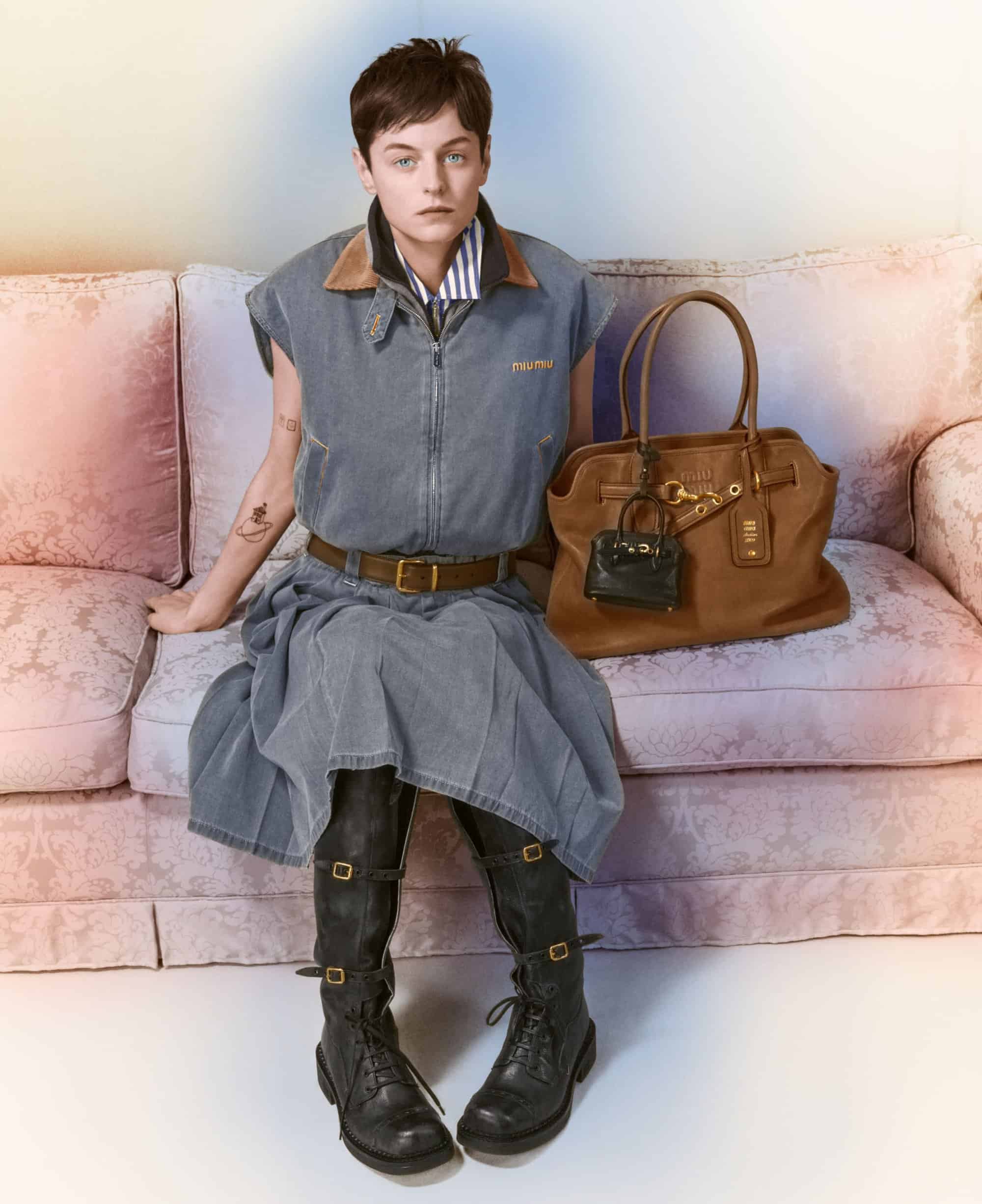 Emma Corrin seated on a patterned sofa wearing grey denim sleeveless top, matching skirt, and black buckled boots with a brown leather handbag