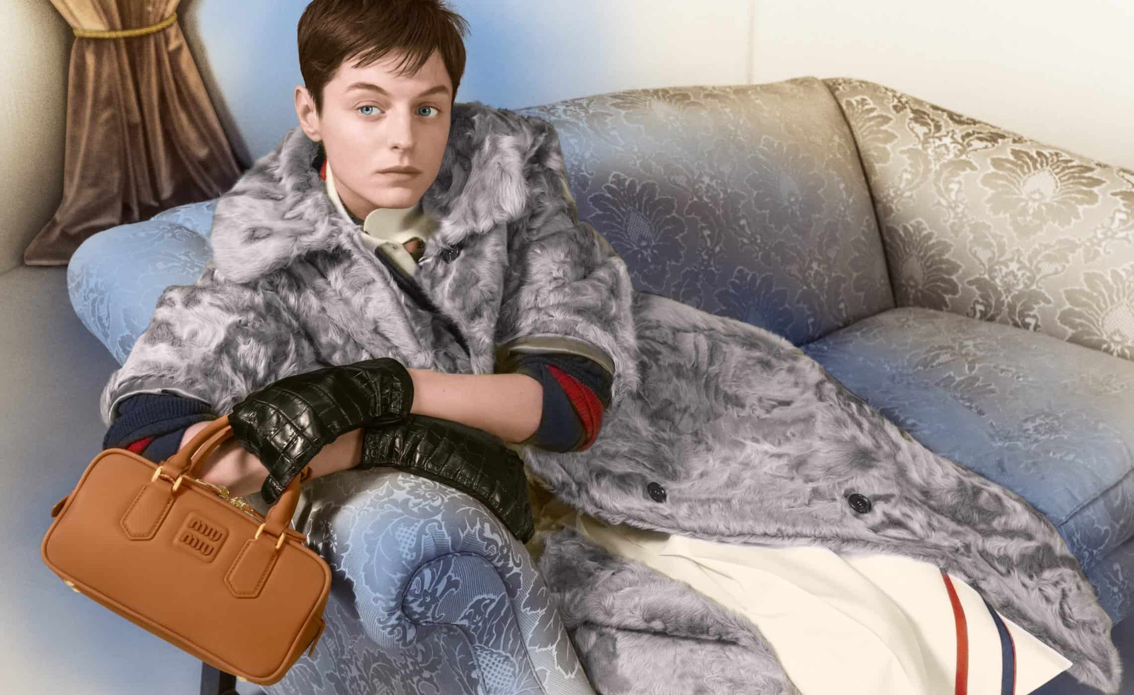 Emma Corrin wearing a grey faux fur coat and leather gloves holding a brown handbag on a patterned sofa for Miu Miu Holiday 2024