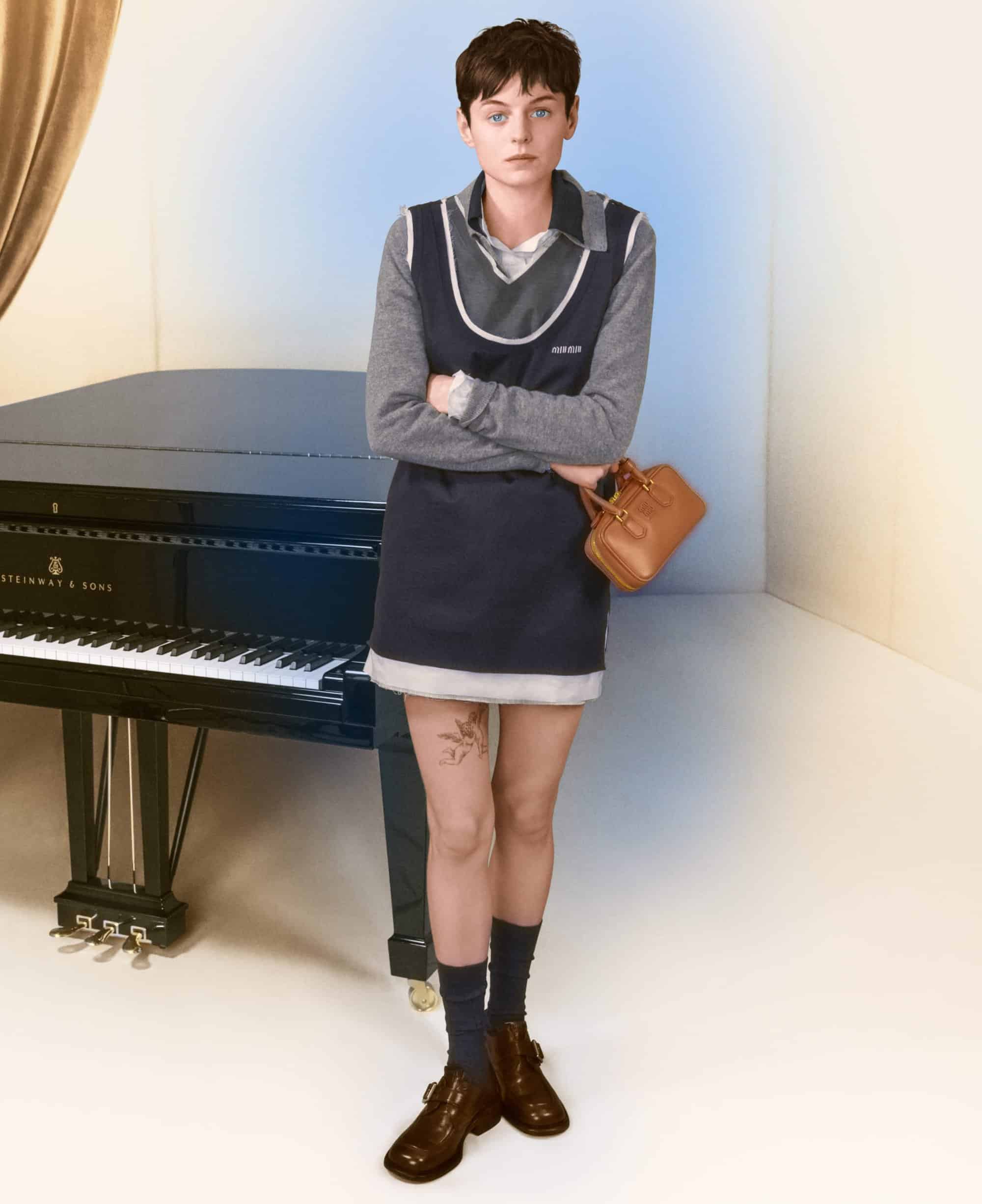 Emma Corrin wearing a layered grey top and navy dress standing beside a black grand piano with a tan handbag for Miu Miu