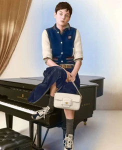 Emma Corrin seated on a black grand piano wearing a blue varsity jacket and holding a white leather handbag