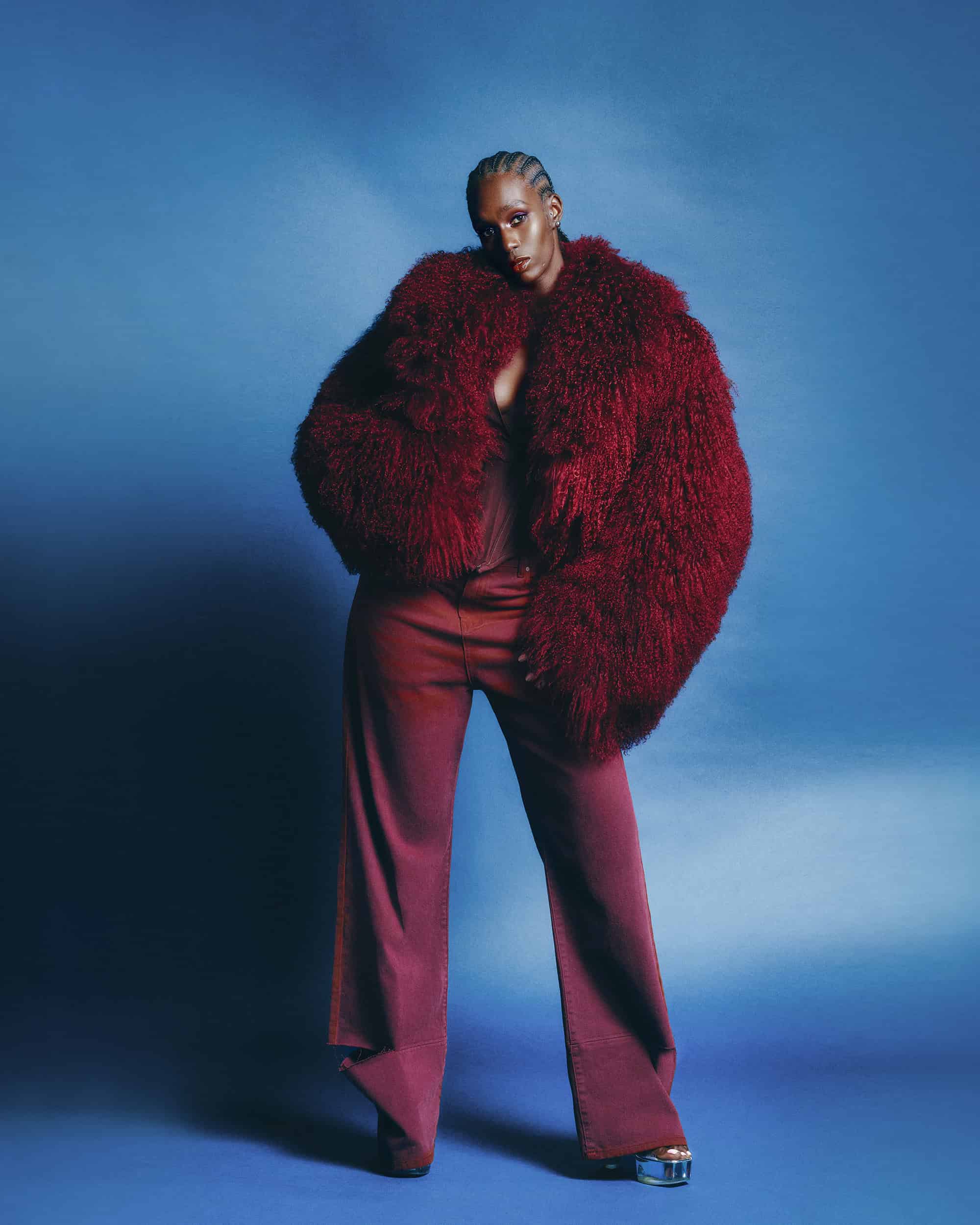 Black woman wearing a voluminous burgundy fur coat and matching trousers against a blue background