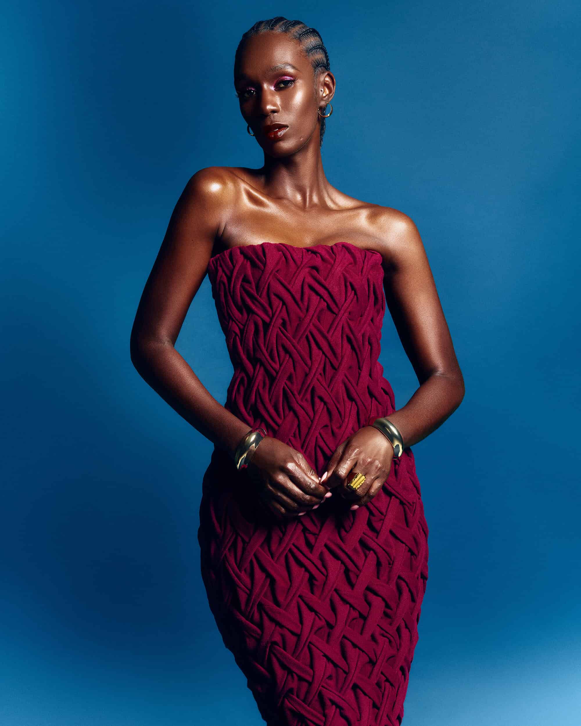 Black woman wearing a strapless textured maroon dress and metallic bracelets against a solid blue background