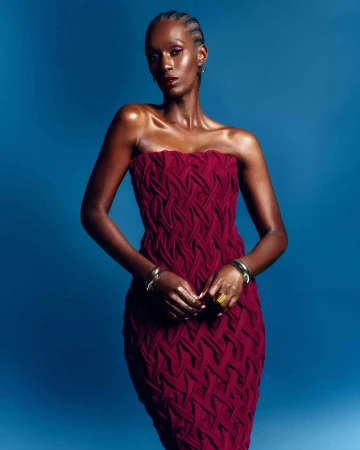 Black woman wearing a strapless textured maroon dress and metallic bracelets against a solid blue background
