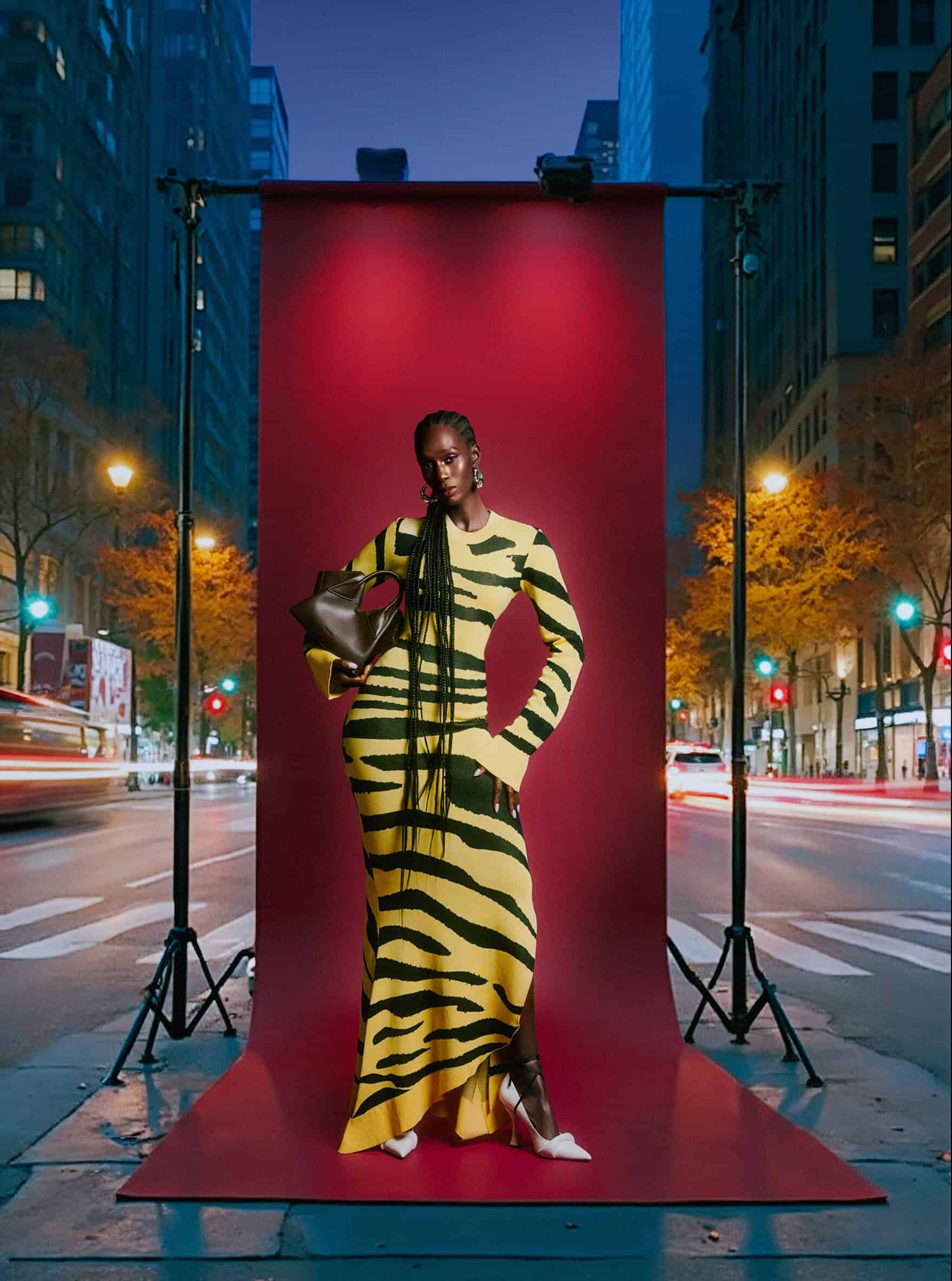 Model in a yellow and black striped dress posing in front of a red backdrop on a city street at night.