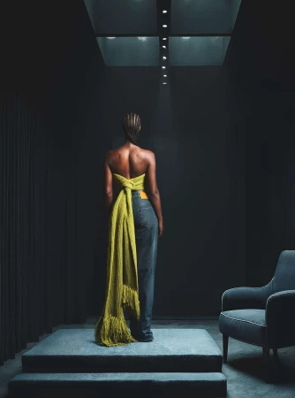 Model seen from behind wearing a green knit top and jeans standing on a raised grey platform under a spotlight