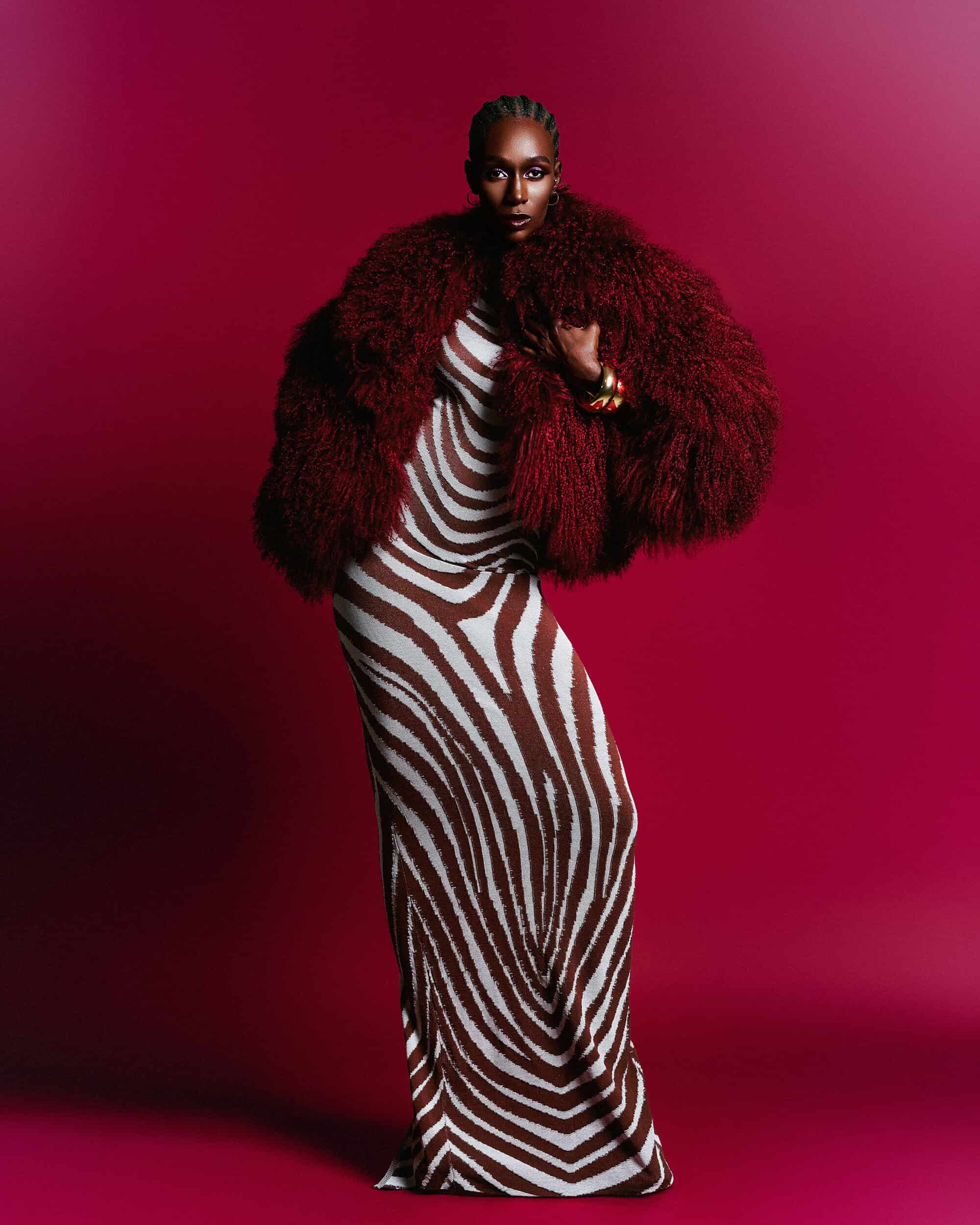 Model in a zebra print dress and textured burgundy fur jacket posing against a magenta gradient studio background.