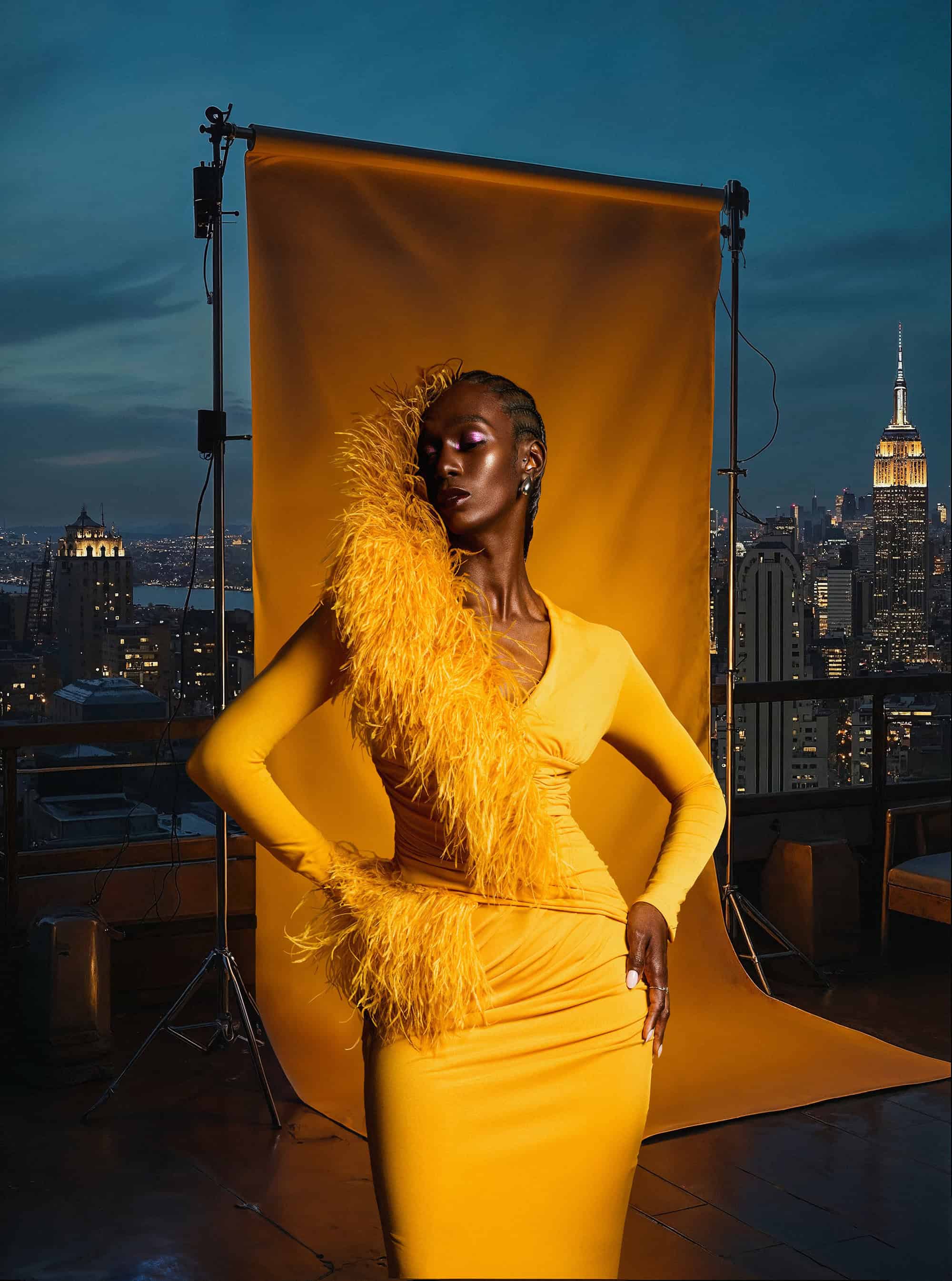 Black woman in a yellow dress with feather trim standing before a mustard studio backdrop on a city rooftop at dusk