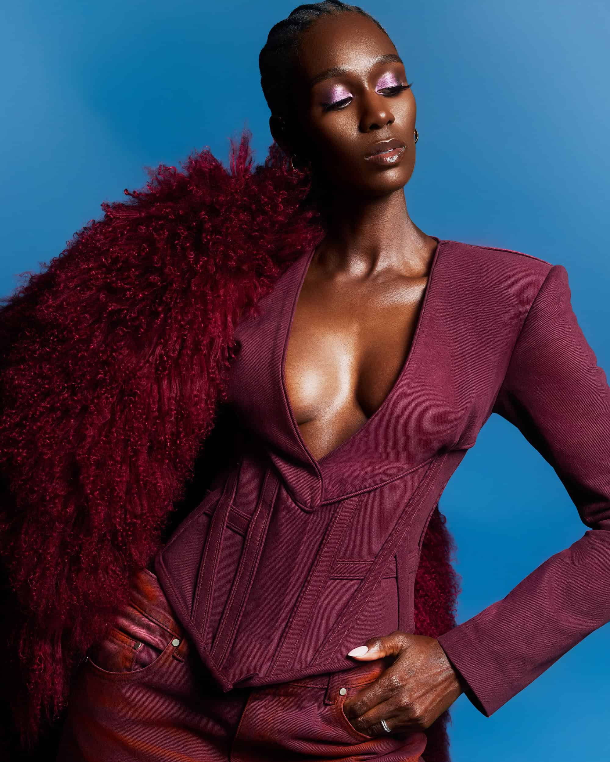 Black woman wearing a structured burgundy blazer and textured red coat against a solid blue background for Hanifa Fall 2024 campaign