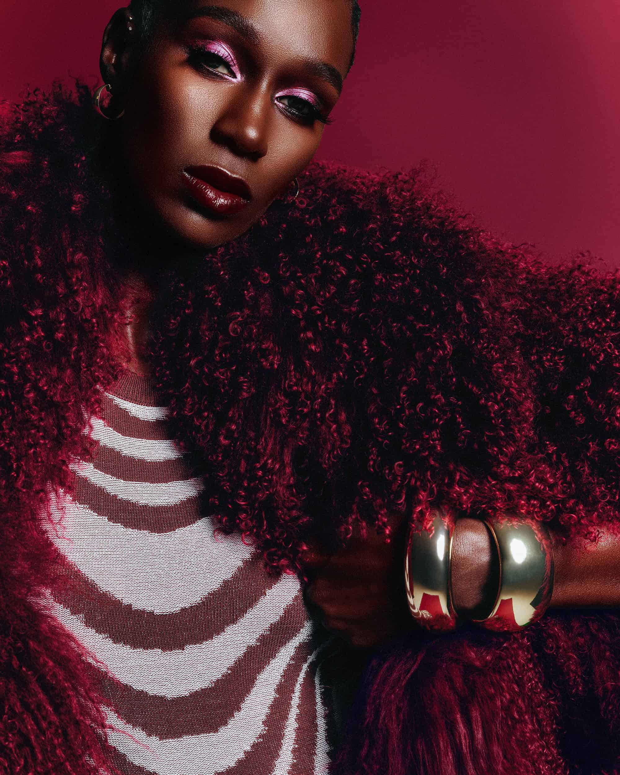 Black woman in a burgundy fluffy coat and striped top, wearing gold bracelets, against a rich burgundy background.