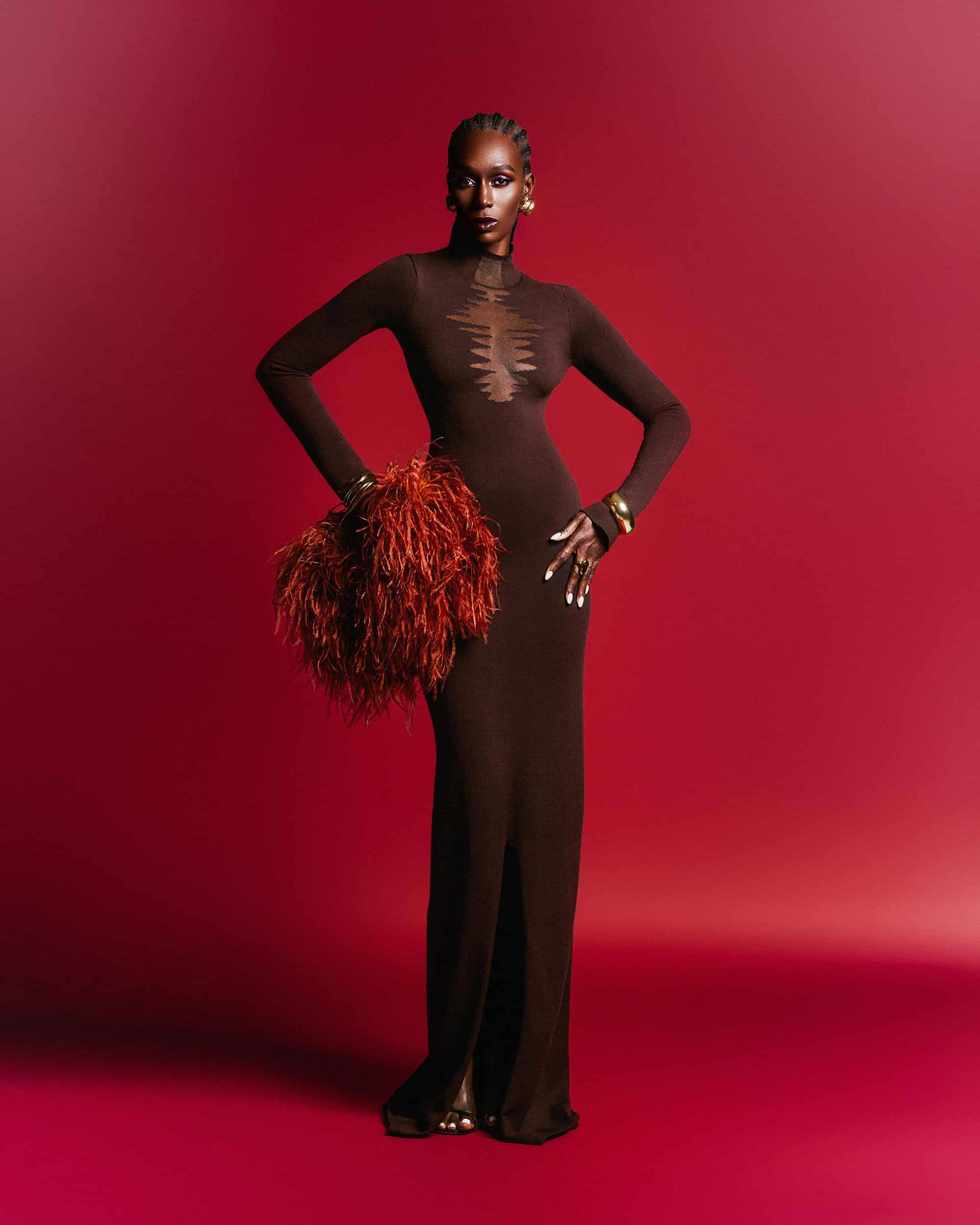 Black woman wearing a long brown gown holding a textured red feather clutch against a solid red studio background.