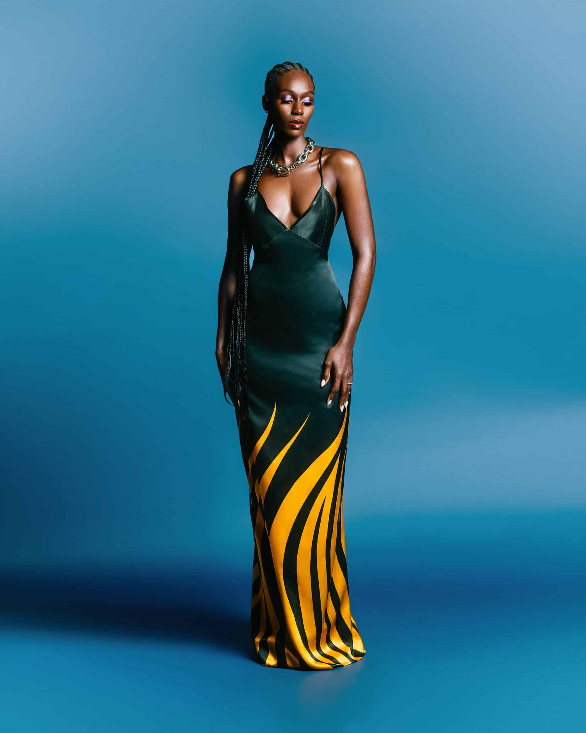 Black woman in a long dark green and yellow patterned slip dress standing against a blue studio background.