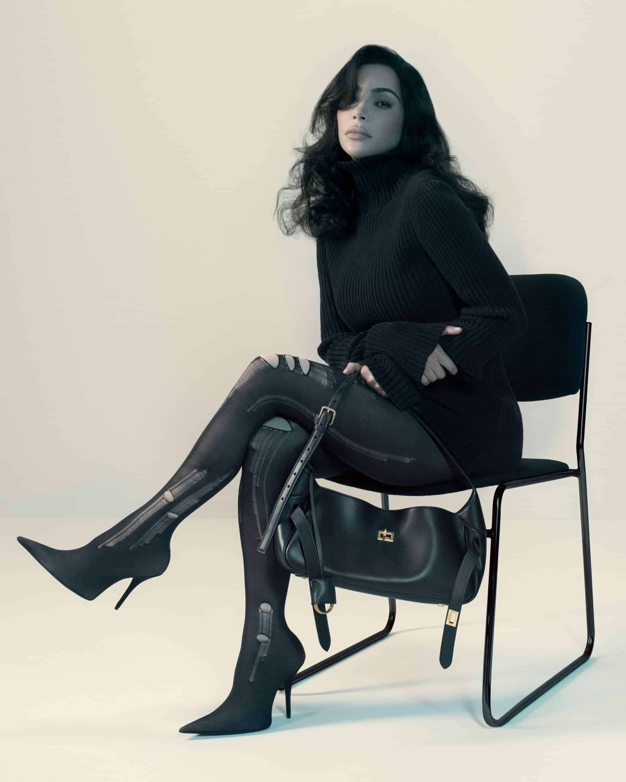 Kim Kardashian seated on a black chair wearing a black turtleneck sweater and black boots, holding a Balenciaga Bel Air bag