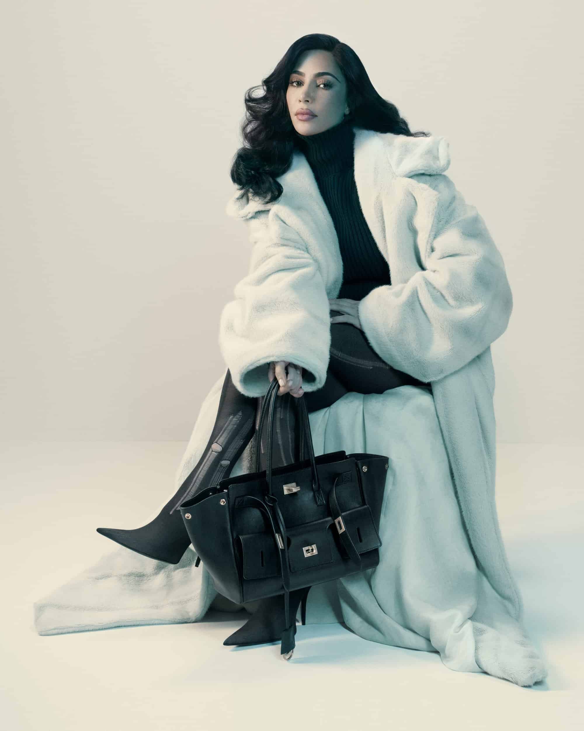 Kim Kardashian in a white faux fur coat holding a black leather handbag against a plain background for Balenciaga